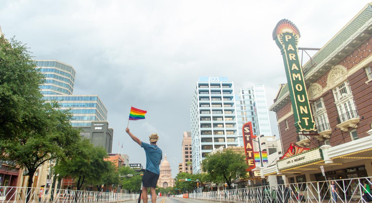 头戴假牛仔帽的男子站在滑板车上,在派拉蒙戏院和国会大厦前挥动着彩虹旗帜 头戴假牛仔帽的男子站在滑板车上,在派拉蒙戏院和国会大厦前挥动着彩虹旗帜